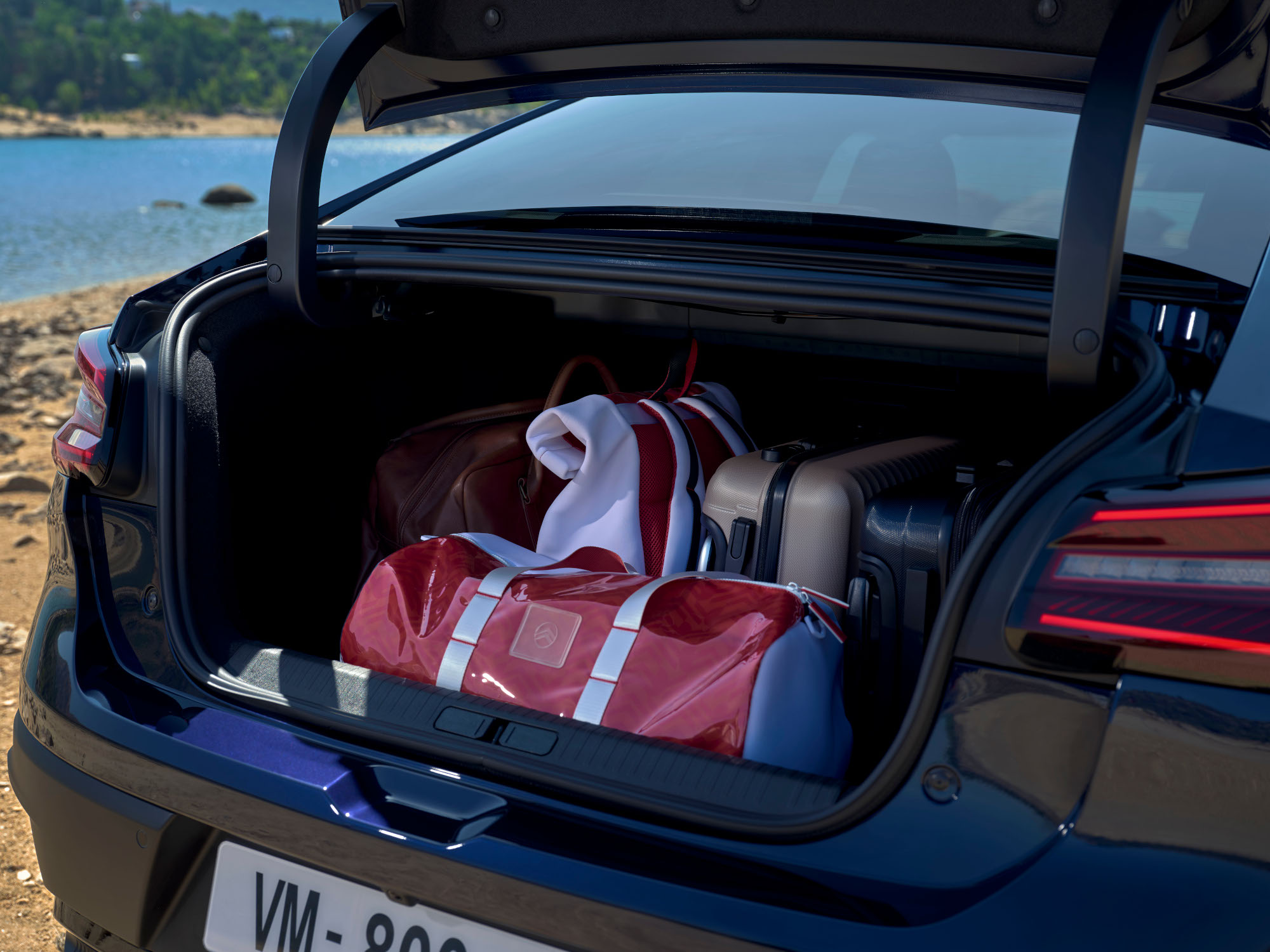 Bagagliaio aperto della Citroën ë-C4 X carico di borse da viaggio vicino a un paesaggio lacustre